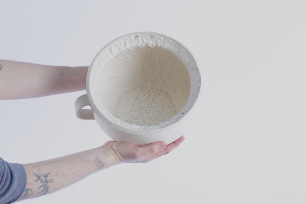 Two hands holding an unfired porcelain chamber pot.