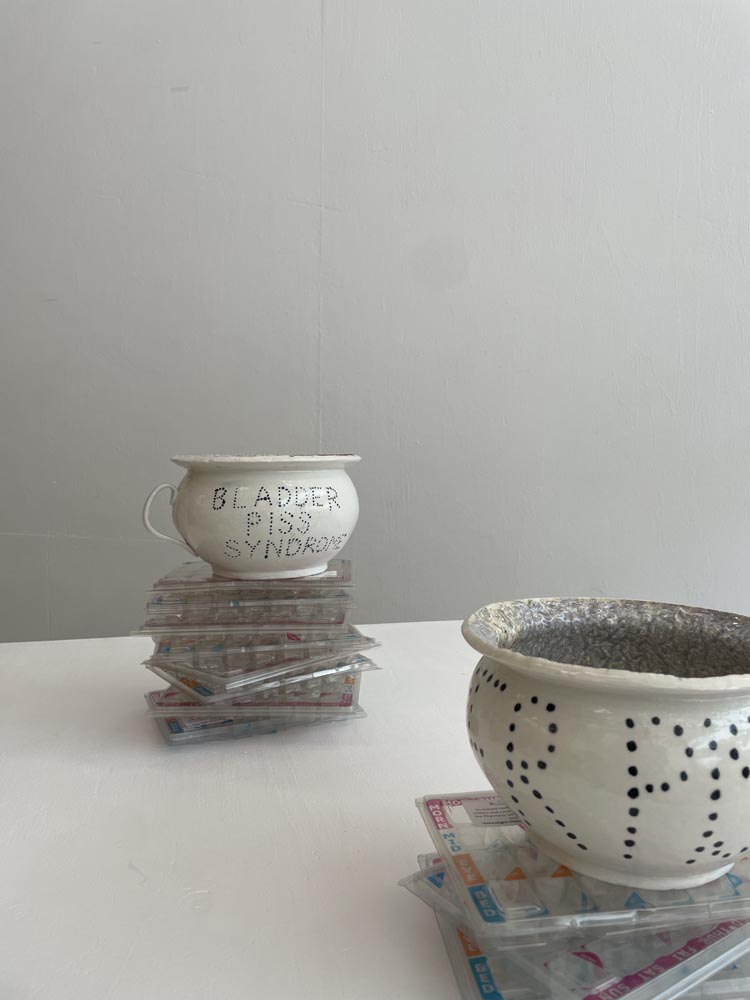 Two porcelain chamber pots perched on piles of dossette boxes on a white surface. One says Bladder Piss Syndrome in dots.