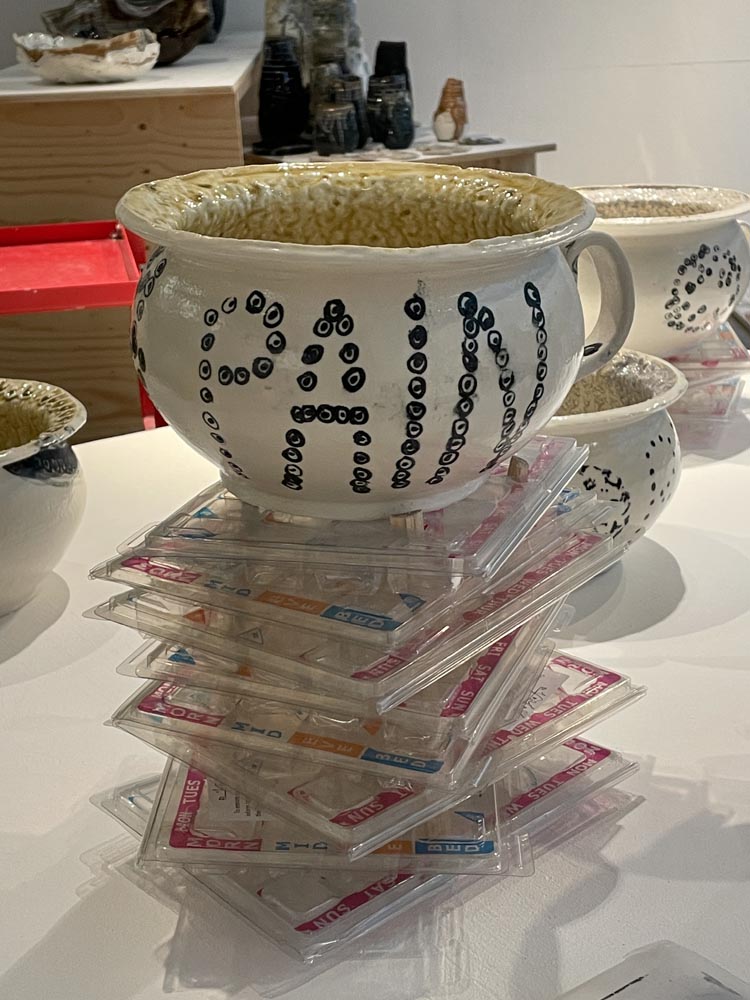 A porcelain chamber pot that says PAIN in capital letters made out of cells. It is on top of a pile of dossette boxes.